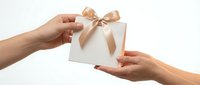 Woman handing wrapped gift box with gold ribbon bow to another person against white background for holiday giving.