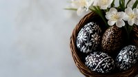 Decorative black and white Easter eggs with traditional folk patterns in woven basket with fresh jasmine flowers, elegant holiday composition on light background.