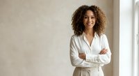 Confident mixed-race businesswoman with curly hair in white blazer smiling with crossed arms in modern office space for corporate presentations.