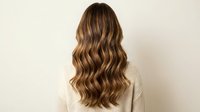 Woman with long wavy brown hair with blonde highlights viewed from behind wearing cream sweater against neutral background for hair salon marketing.