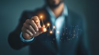 Businessman in dark suit drawing glowing financial chart graph with finger on virtual screen, demonstrating market growth analysis in blue atmosphere.