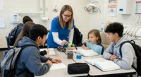 Female teacher with glasses instructing diverse students in modern classroom setting. Educational teamwork and learning environment for academic success.