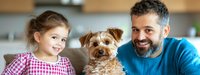 Happy family moment with bearded father, young daughter in checkered dress and Yorkshire terrier puppy relaxing at home, panoramic lifestyle shot showing bonding and joy.