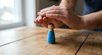 Protective hands covering small blue wooden figure on rustic table symbolizing care, security, insurance and business protection concepts.