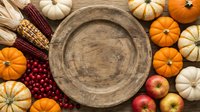 Rustic wooden plate surrounded by autumn harvest vegetables including orange and white pumpkins, corn, apples, and cranberries on wooden surface for fall seasonal designs.