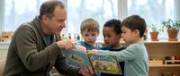 Adult male educator reading storybook with diverse group of young children in bright classroom setting with plants and windows, promoting literacy and early childhood learning.
