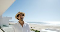 Man in straw hat and white shirt smiling on modern beach house balcony overlooking ocean and sandy shore on sunny day.