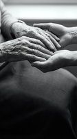 Elderly wrinkled hands gently held by younger smooth hands in black and white showing intergenerational care, support and compassion between family members.