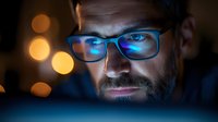 Focused businessman with glasses working late at computer screen with blue light reflection and warm bokeh background for technology concepts.