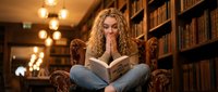 Young blonde woman with curly hair reading book in cozy library armchair surrounded by bookshelves with warm lighting for education and literature concepts.
