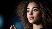 Young African American woman with curly hair and glowing skin gazing upward with contemplative expression against dark background.