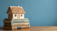 Wooden house model on stack of architecture and homebuilding books against blue background for real estate education and construction learning concepts.