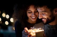 Young mixed race couple smiling at glowing gift box with magical light bokeh background creating romantic holiday atmosphere at night.