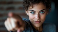 Young mixed race woman with bright blue eyes and curly dark hair smiling confidently while pointing finger at camera against blurred background.