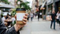 Person holding takeaway coffee cup with blank label on urban street with blurred pedestrians and storefronts in background for beverage branding and cafe marketing.