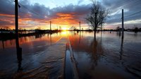 Dramatic sunset over flooded street with utility poles and bare trees reflected in still water, creating mirror effect against fiery orange and purple sky.