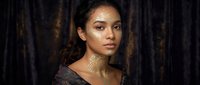 Young mixed-race woman with golden face paint and braided hair against dark textured background for beauty and fashion concepts.
