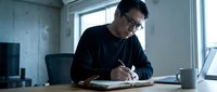 Man with dark hair and glasses, wearing dark casual clothing, writing in notebook at desk in modern office space with laptop and coffee mug, focused on work.