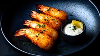 Crispy golden fried shrimp tempura served on black plate with creamy dipping sauce and lemon. Garnished with fresh herbs, dramatic dark food photography.