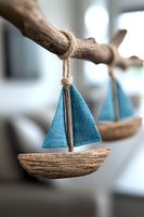 Handcrafted driftwood sailboat ornament with turquoise blue fabric sail and natural twine hanging from wooden branch, coastal home decor against blurred background.