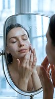 Woman with dark hair and light skin tone touching her face while looking at mirror reflection in bathroom, skincare routine and beauty treatment concept.