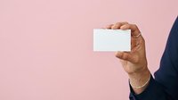 Hand of a mature man with medium brown skin tone wearing a dark navy blazer, holding a blank white business card against a soft pink background, suitable for corporate branding and business presentations.