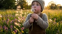 Young child with light skin tone and blonde hair wearing a knit beanie and brown vest, holding a dandelion in a sunlit meadow filled with wildflowers during golden hour.