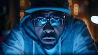 African American man in blue hoodie and glasses illuminated by screen light at night, focused expression showing digital engagement.