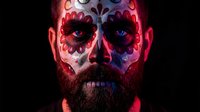 Bearded man with colorful Day of the Dead sugar skull face paint featuring red and blue decorative patterns against dark background for Halloween celebrations.