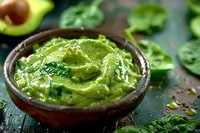 Fresh guacamole dip with mashed avocado and herbs in rustic ceramic bowl on dark background, surrounded by spinach leaves and ingredients.