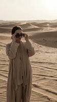 Woman in beige flowing garment adjusting oversized sunglasses in vast desert landscape with rolling sand dunes. Fashion and travel photography.