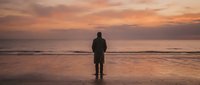 Silhouetted male surfer standing on wet sand facing ocean at golden hour sunset, holding surfboard, contemplative coastal scene for travel and lifestyle content.