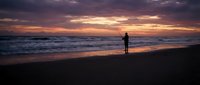 Silhouette of person fishing on ocean beach at dramatic sunset with colorful sky and waves rolling onto shore creating peaceful coastal scene.