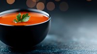 Fresh tomato soup garnished with parsley leaf in black ceramic bowl against dark background with bokeh lights, minimalist food photography composition.