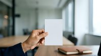 Male hand holding blank white card in modern office workspace with natural light and wooden desk for business branding mockup.