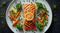 Grilled salmon fillet with lemon slice served with fresh vegetables including broccoli, carrots, peas, and corn on white plate.
