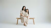 Asian mother and daughter in matching beige outfits sitting on wooden bench against white background, family portrait studio photography for lifestyle content.