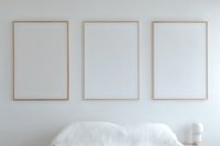 Three empty rose gold frames mounted on white wall above white bedding, creating minimalist bedroom interior design with modern aesthetic and clean lines.