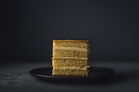 Layered honey cake slice on dark plate against moody black background, showcasing golden sponge layers with cream filling for bakery and dessert concepts.