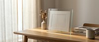 Minimalist wooden desk workspace with empty white picture frame, ceramic vase with dried branches, coffee mug and books in soft natural lighting.
