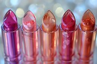 Colorful lipsticks in shades of pink, coral, and red with shimmering texture displayed in a row against blurred bokeh background.