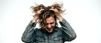 Young man with long wavy brown hair and Caucasian ethnicity wearing denim jacket, hands in hair, expressive pose for lifestyle and fashion content.