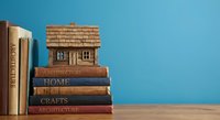 Miniature wooden house model sitting on stack of architecture and home design books against blue background for real estate education concepts.