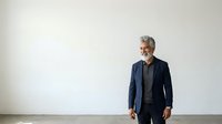 Mature man with gray hair and beard, Caucasian ethnicity, wearing dark navy blazer and black shirt, standing against white wall in professional business setting.