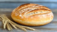 Fresh baked crusty sourdough bread loaf with golden brown crust and wheat ears on rustic wooden background, close up view of artisanal bakery product.