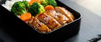 Glazed chicken teriyaki with steamed broccoli and carrots in black bento box on wooden surface for healthy meal prep and Japanese cuisine presentation.