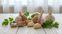 Fresh garlic bulbs and cloves with green parsley leaves arranged on white wooden table for cooking and culinary preparations in kitchen setting.