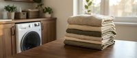 Stack of neatly folded clean towels on wooden table in modern laundry room with washing machine and natural light from window.