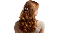 Young woman with long wavy copper hair styled with butterfly clip shown from behind, demonstrating professional hairstyling on light background for beauty and fashion content.