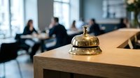Golden brass service bell on wooden reception desk in modern hotel lobby with blurred business people in background for hospitality industry.